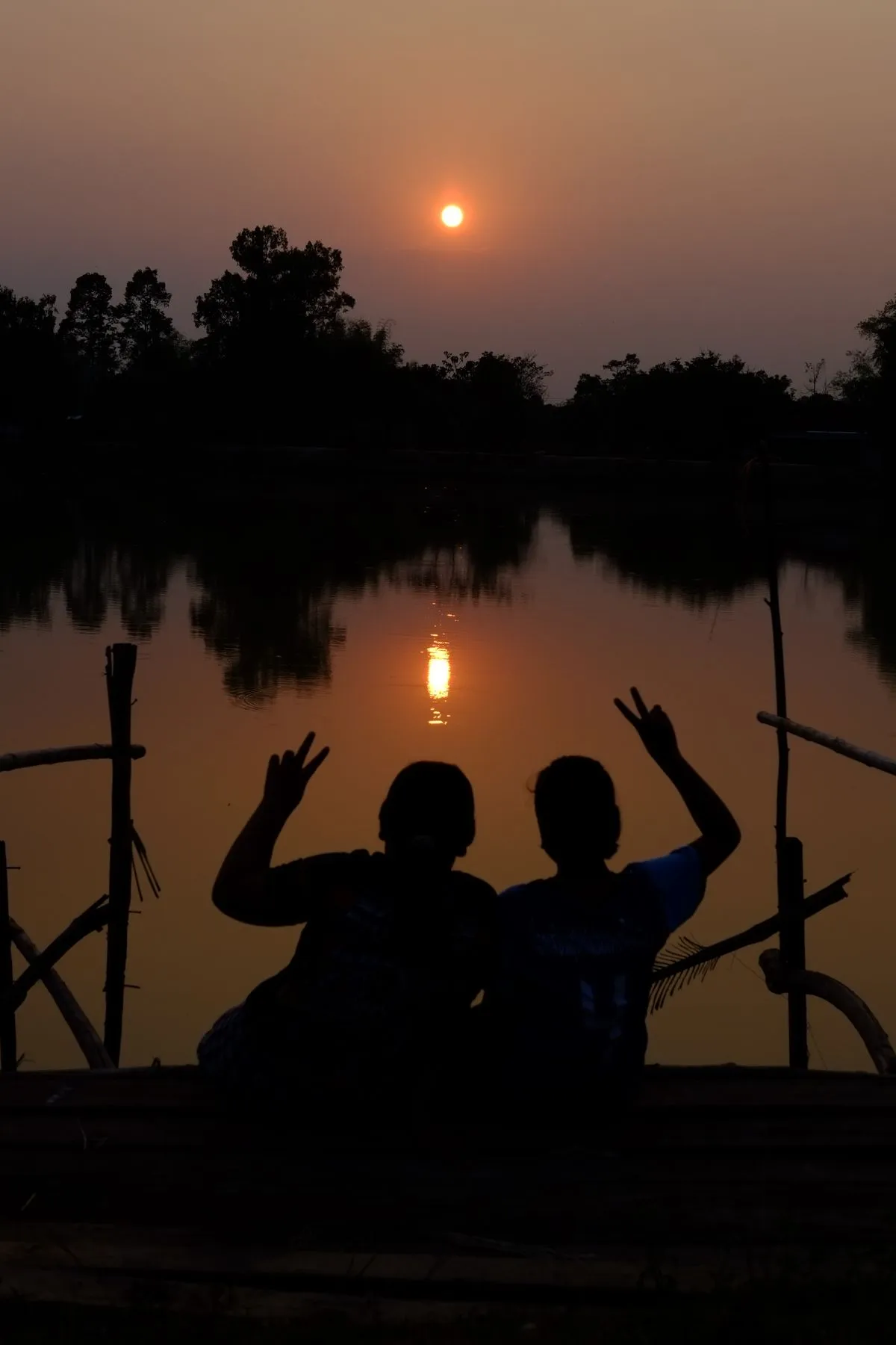 Two friends sitting together in silhouette by a lakeshore at sunset
