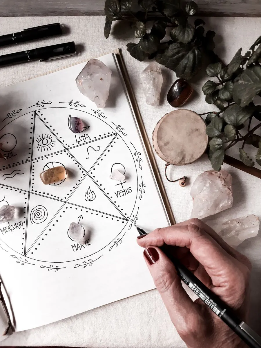 A hand drawing an astrology chart surrounded by crystals and plants